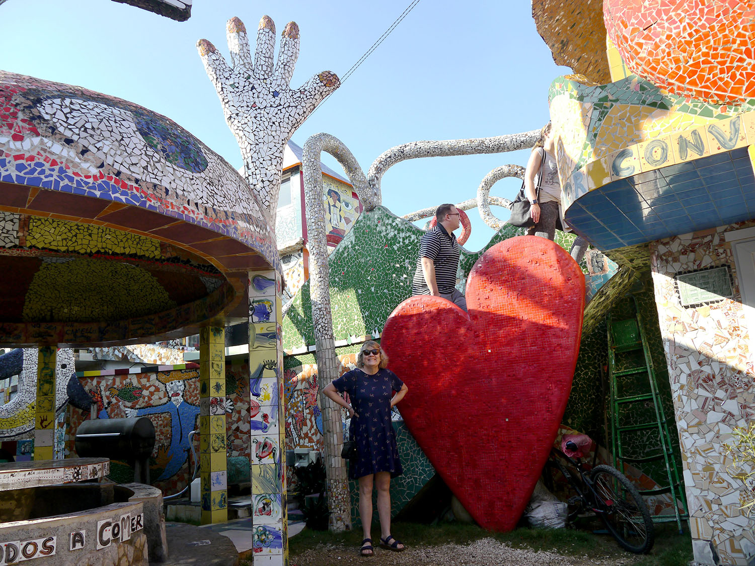 A woman stands in front a giant red heart made out of mosaics, while a man climbs up stairs of mosaics behind her.