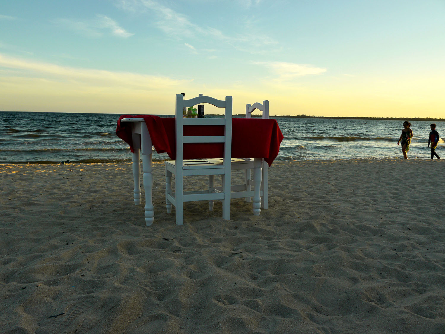 A white table with a red table cloth on the sand on the beach.
