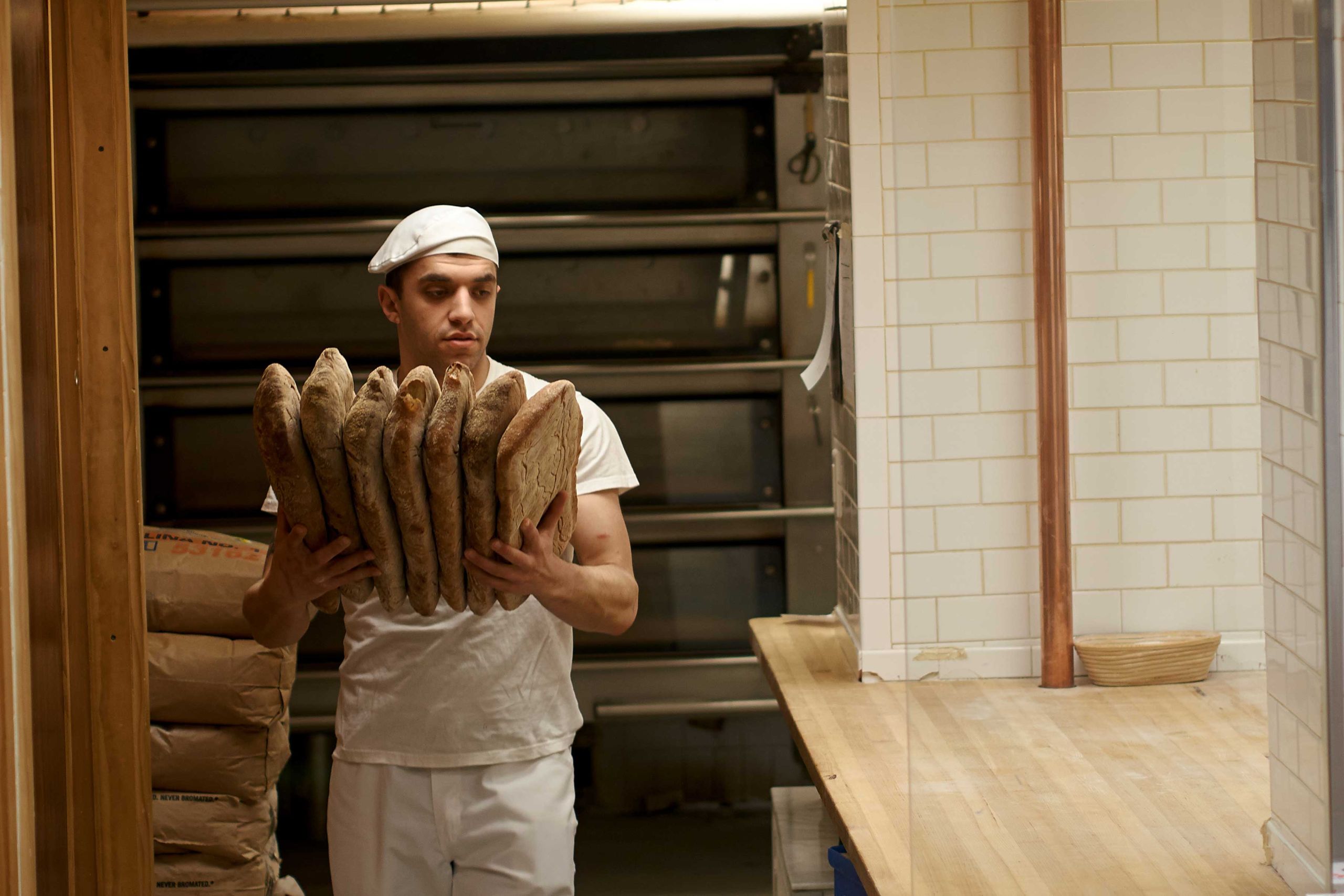 A baker walks from the oven carrying loaves of bread.