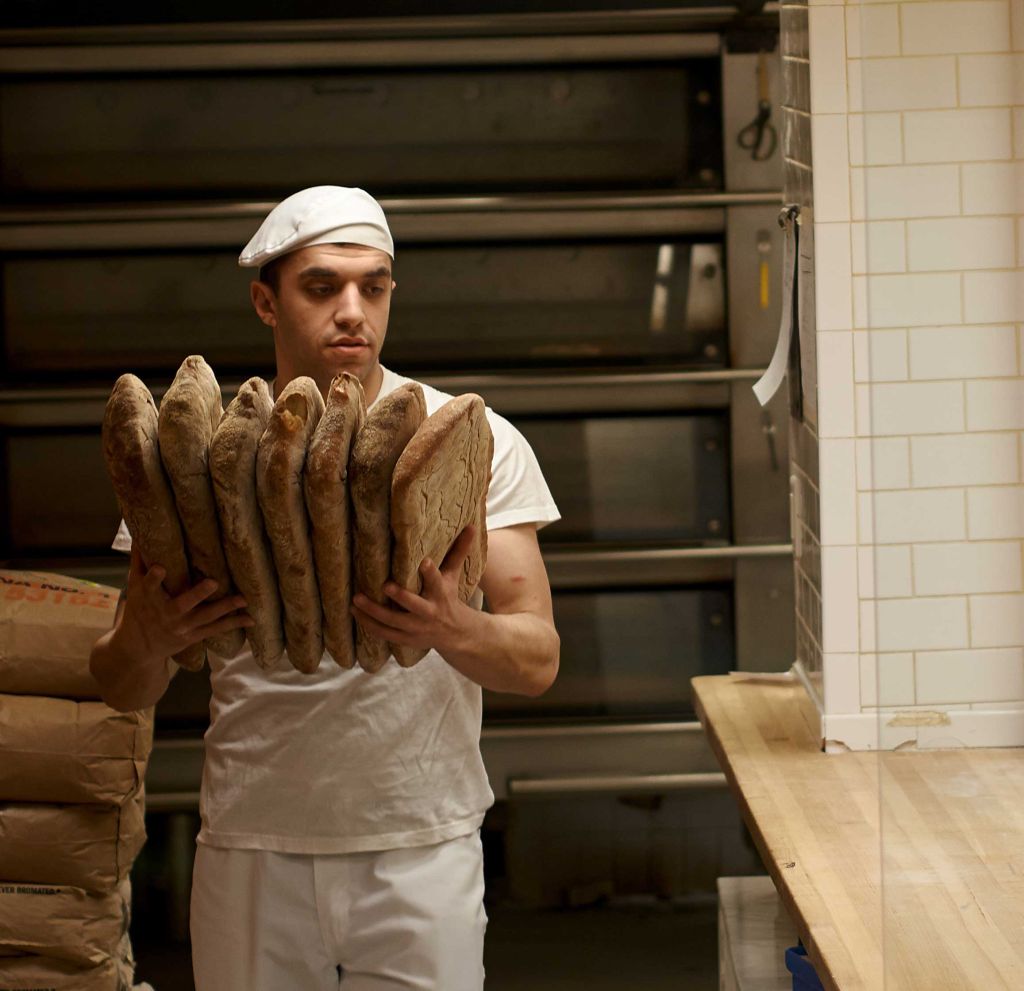 A baker walks from the oven carrying many loaves of bread.