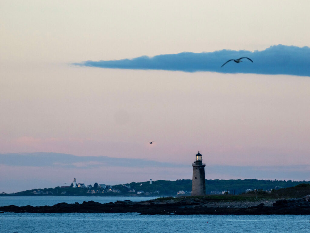A lighthouse on a rocky patch of land in the blue ocean at sunrise.