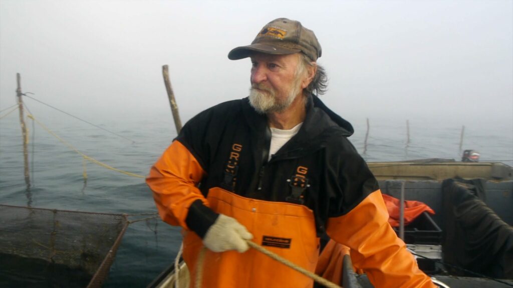 A fisherman with a beard in orange overalls on a boat.