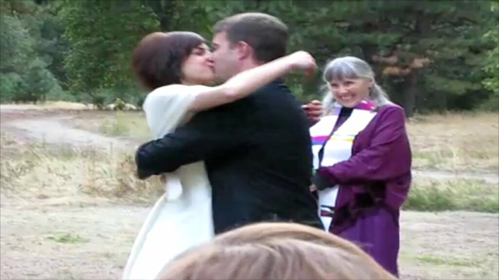 A bride and groom kiss while a female pastor smiles in the background.
